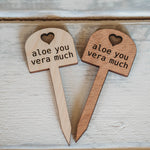 Two wooden plant markers with a heart-shaped top, both inscribed with the phrase 'aloe you vera much', are placed on a light wooden textured surface with a slight rustic appearance.