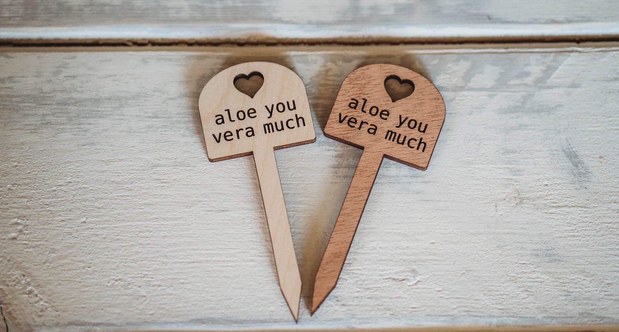 Two wooden plant markers with a heart-shaped top, both inscribed with the phrase 'aloe you vera much', are placed on a light wooden textured surface with a slight rustic appearance.