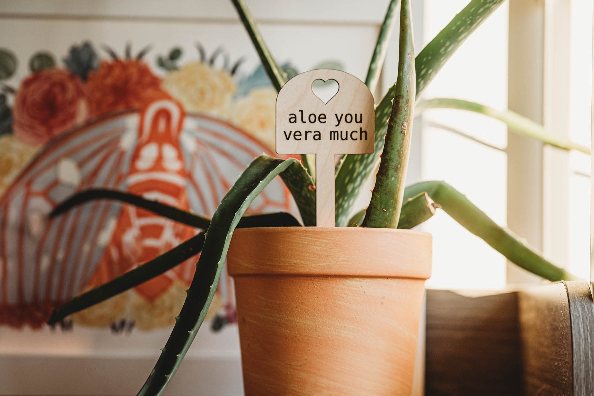 An aloe plant in a terracotta pot with a wooden marker reading 'aloe you vera much', placed on a windowsill. Behind it is a decorated framed artwork with a red and orange pattern